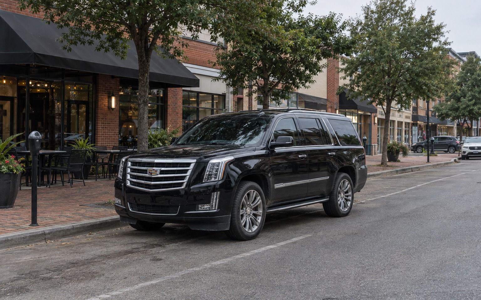 Black Cadillac Escalade parked by a restaurant patio on a tree-lined street